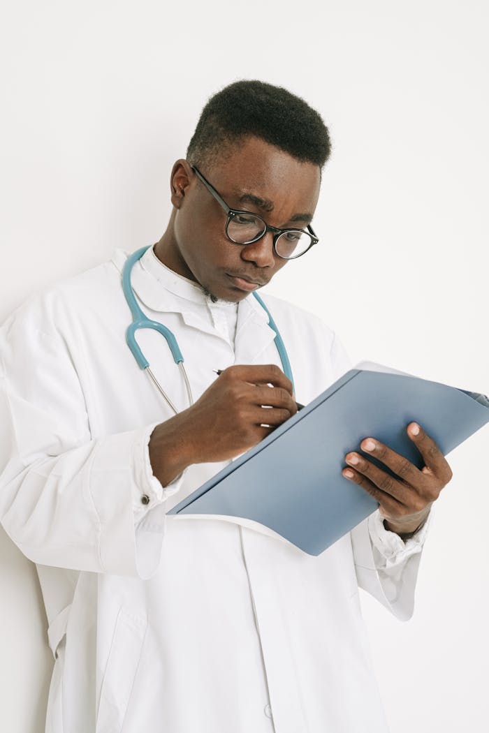 A doctor wearing eyeglasses writes on a medical chart indoors, displaying professional focus.
